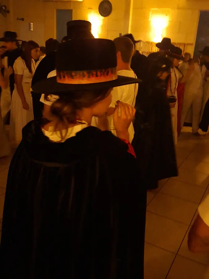 Pessoa paramentada com capa e chapéu pretos, de costas, sob luz baixa.